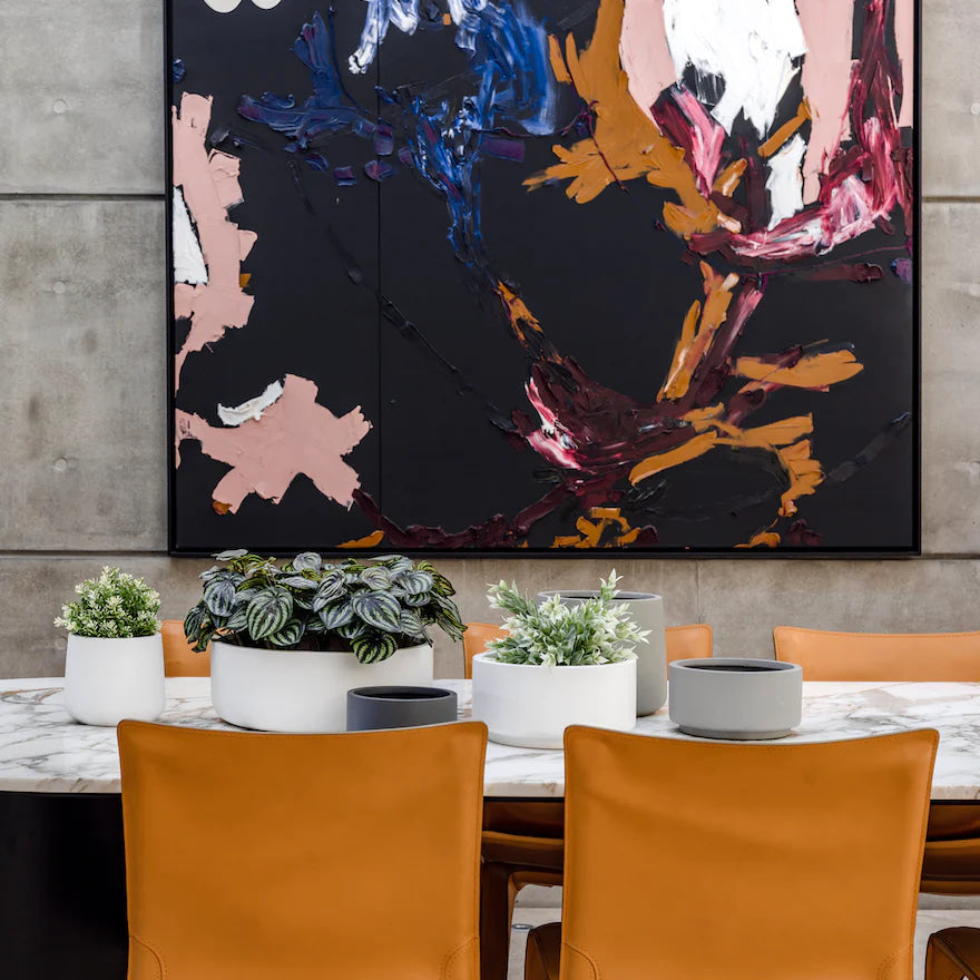 Elegant indoor plant arrangement featuring a variety of The Balcony Garden pots on a marble table, complemented by tan leather chairs and a vibrant abstract painting in the background
